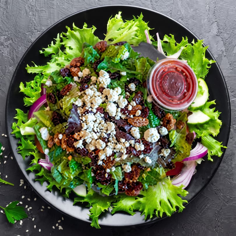 Overhead view of a vibrant green salad with blue cheese, pecans, dried cranberries, and red onion.