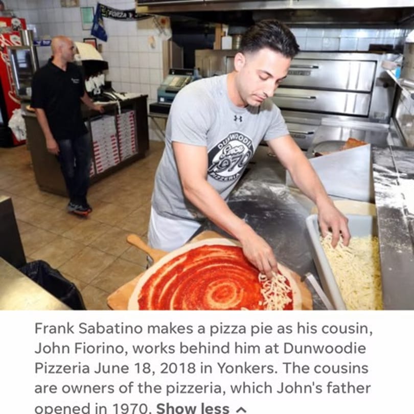 A man in a gray shirt making a pizza with sauce and cheese, another man in the background.