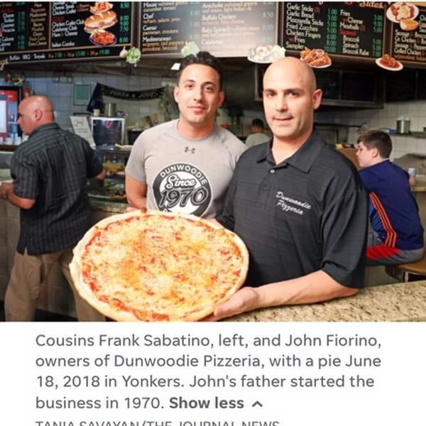 Two men, owners of Dunwoodie Pizzeria, proudly hold a large cheese pizza in their restaurant.